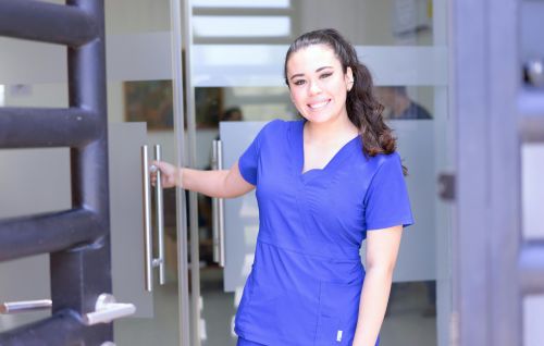 Smiling female nurse in blue scrubs opens a glass door in a healthcare setting.